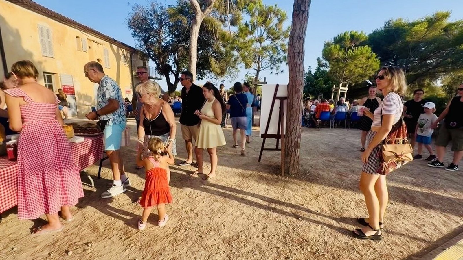 Families gathered in a village square at golden hour, a checkered-cloth stall and a crowd seated under the pines