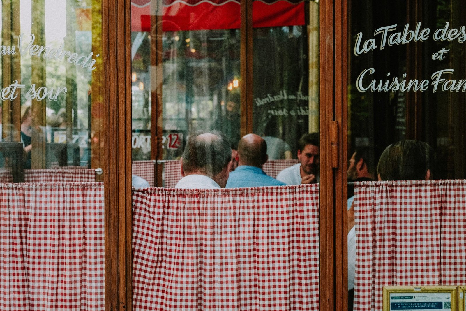 A bistro's gingham-curtained window at dusk, "la Table des Amis, Cuisine Familiale" painted on the glass, a table of regulars talking inside