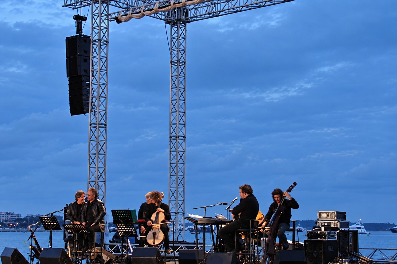 A chamber ensemble playing cello and double bass on an open-air stage at dusk, the bay behind them